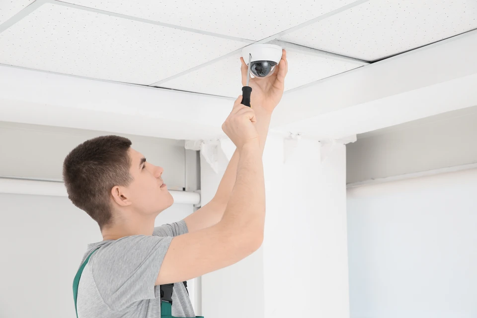 Jeune homme technicien homme installant caméra de surveillance CCTV avec un tournevis sur mur blanc debout à l'intérieur